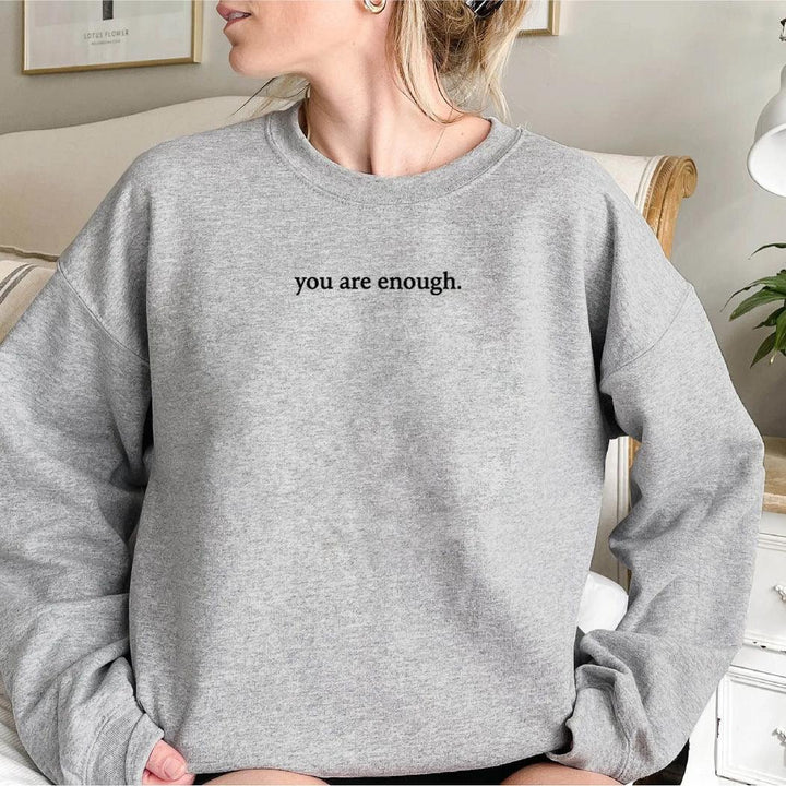 woman wearing light gray sweatshirt with black "you are enough" text in a cozy living room