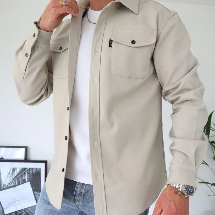 Man wearing beige button-up shirt jacket over white t-shirt with jeans, silver watch, and rings