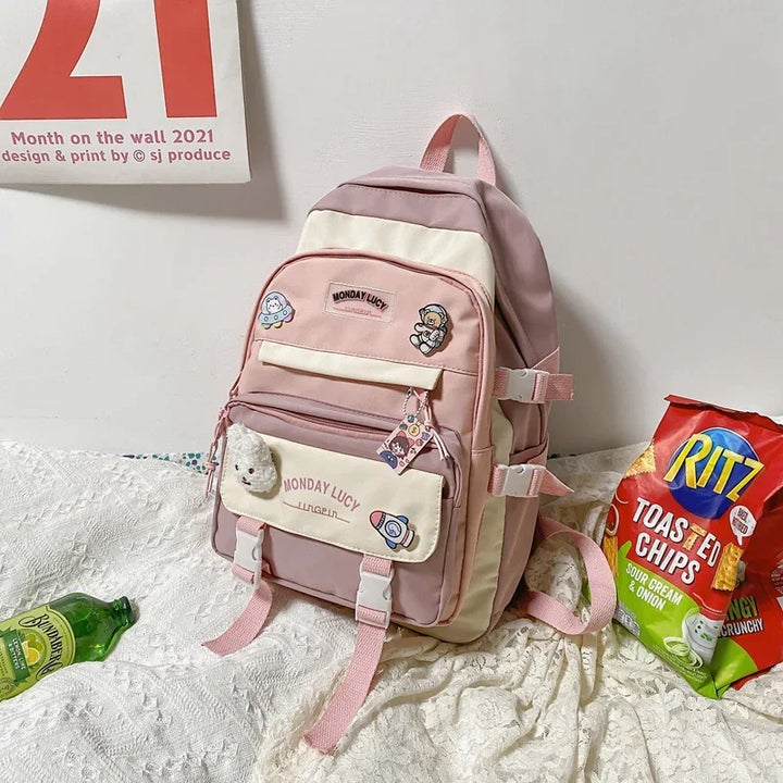 Pink and cream Monday Lucy backpack with cartoon patches on white textured fabric beside Ritz toasted chips and green bottle