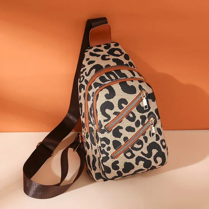 Leopard print sling bag with brown leather trim and black adjustable strap on beige surface