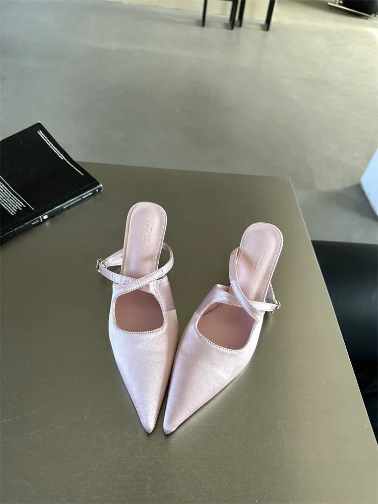 Pair of light pink satin pointed-toe mule heels with ankle straps on a green table indoors