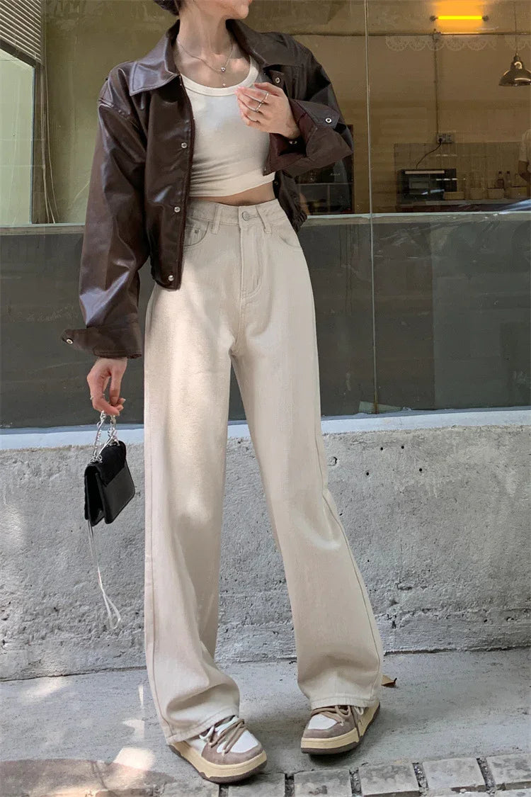 Woman wearing cropped brown leather jacket, white tank top, beige wide-leg pants, and sneakers holding small black purse
