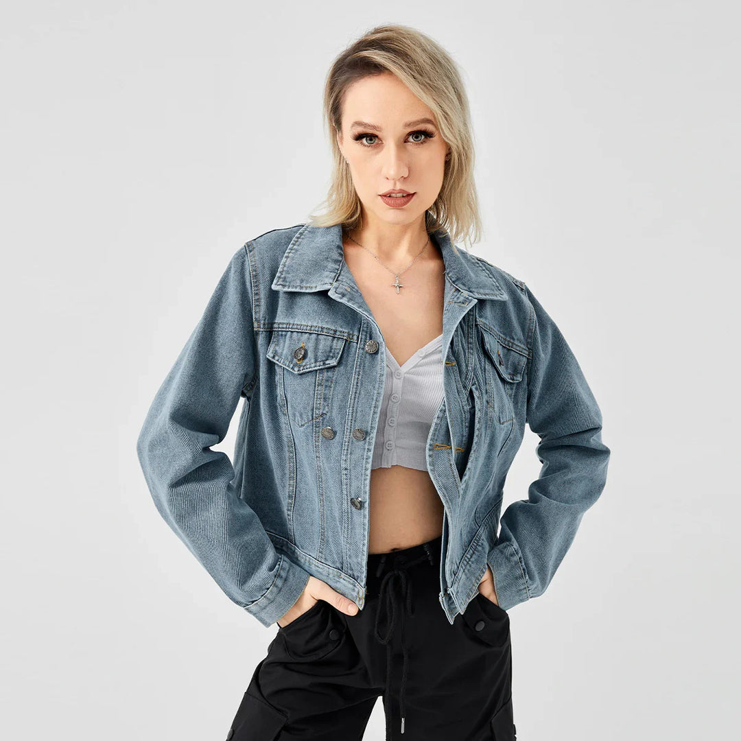 Blonde woman wearing light blue denim jacket, white cropped button top, and black pants posing against gray background