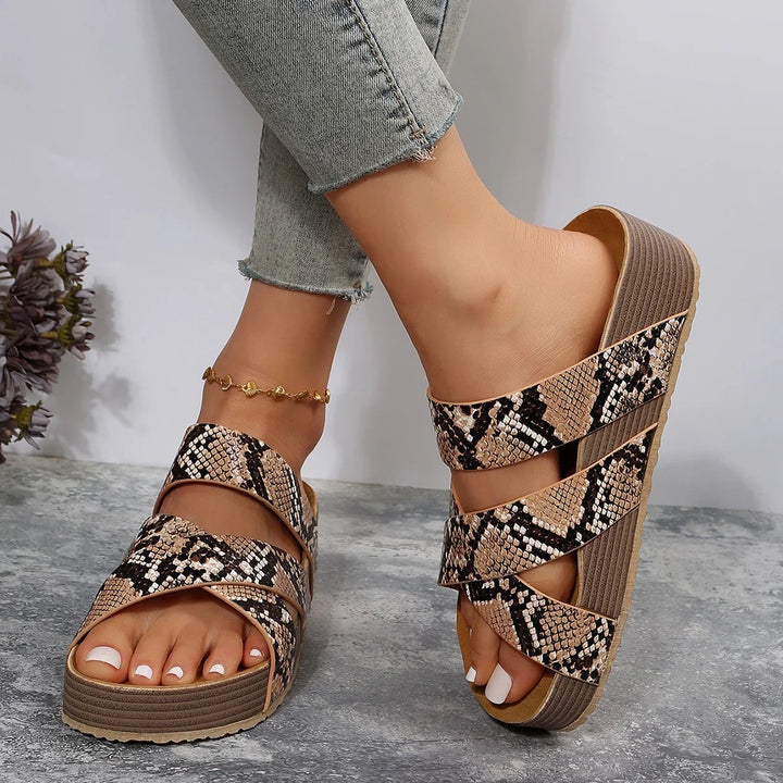 Feet wearing snake print platform sandals with crisscross straps and gold anklet on gray stone floor