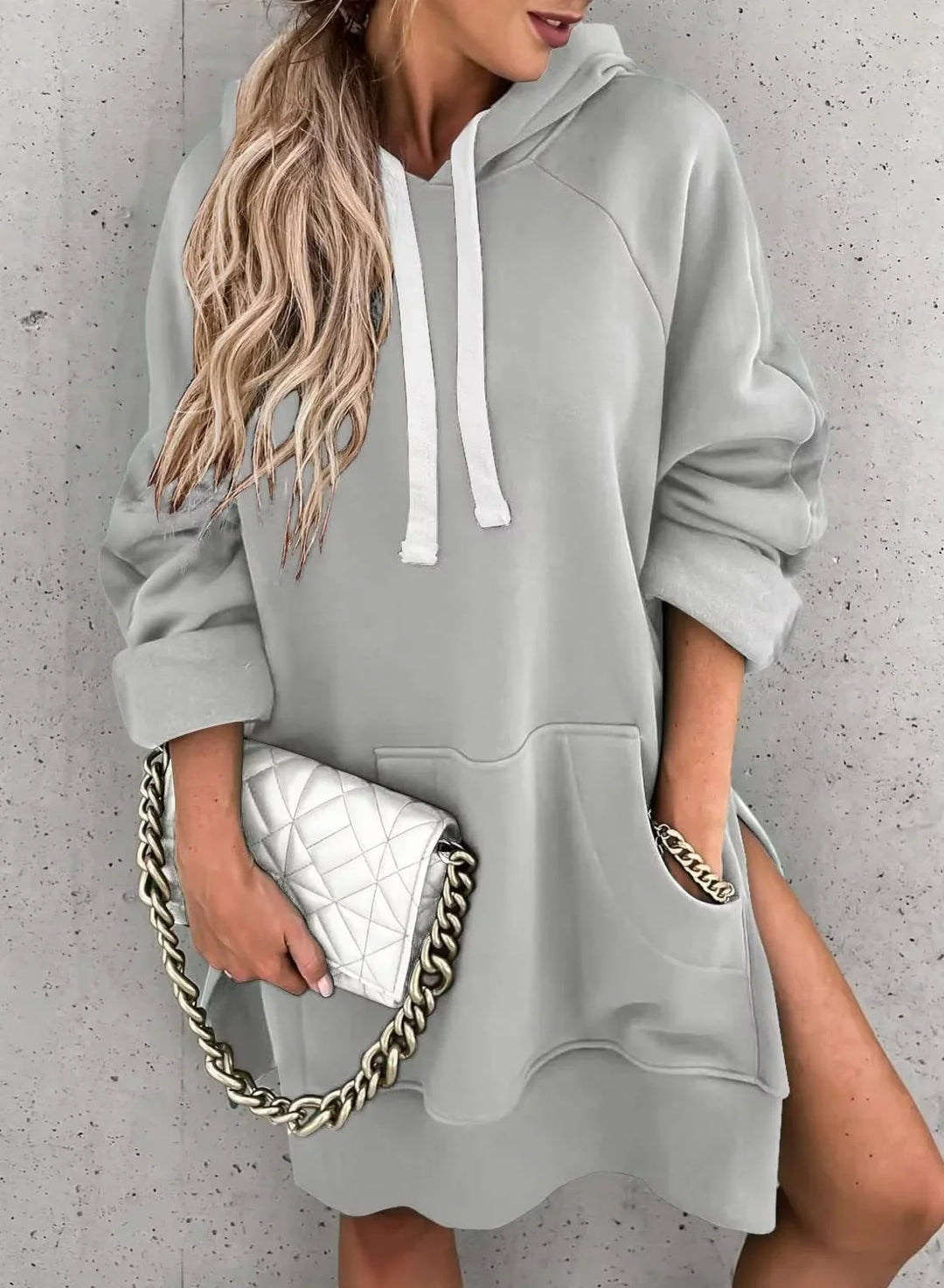 Woman in light gray hoodie dress holding white quilted purse with gold chain against concrete wall