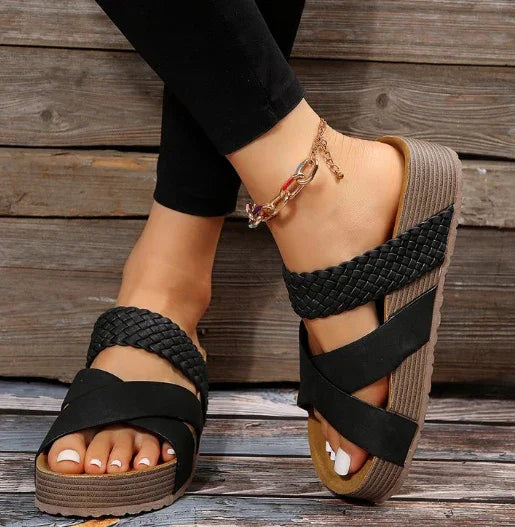 Black woven strap platform sandals on feet with black leggings and gold chain anklet on wooden background