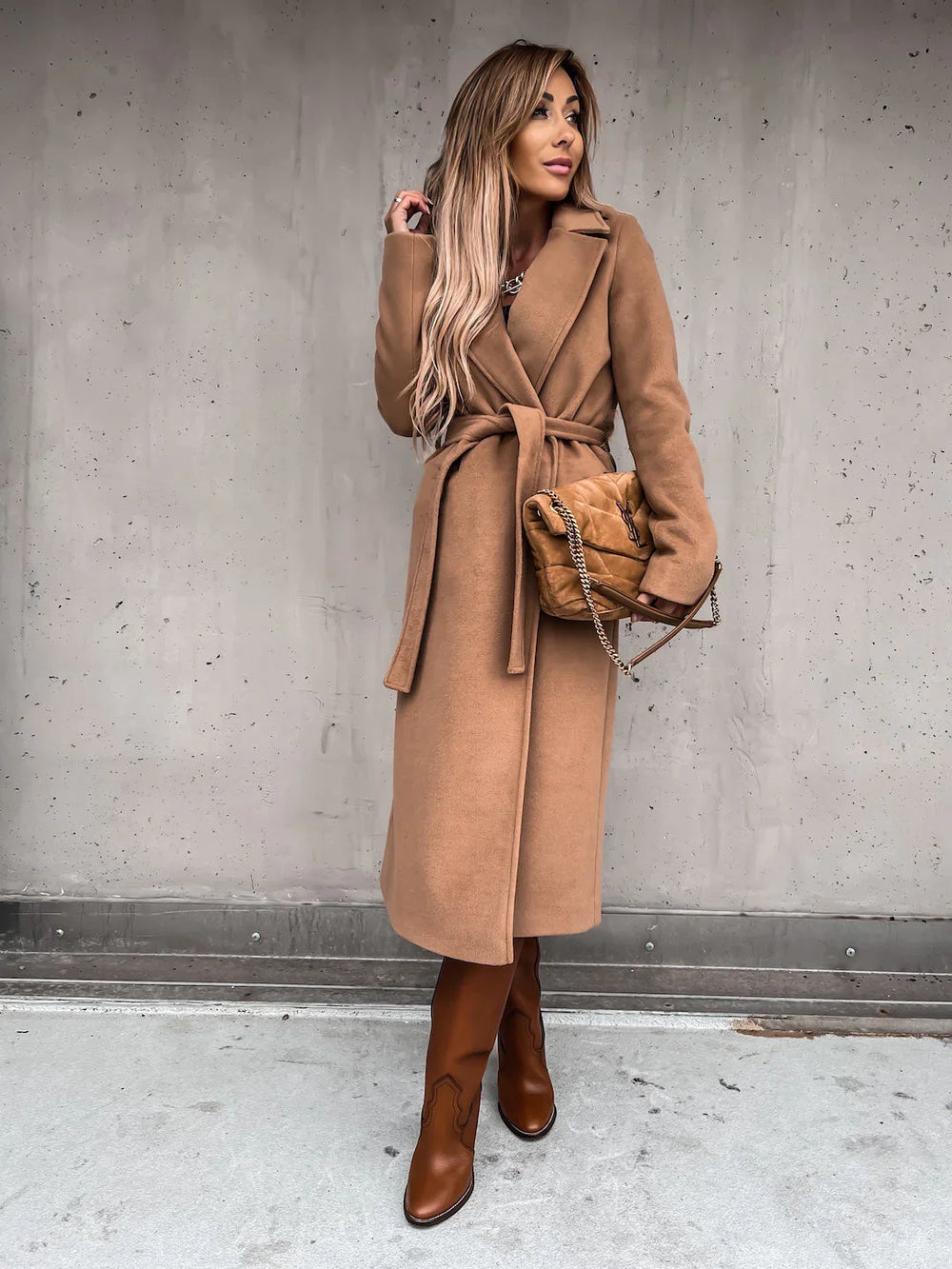 Woman in long camel coat and brown leather boots holding a tan quilted chain purse against gray wall