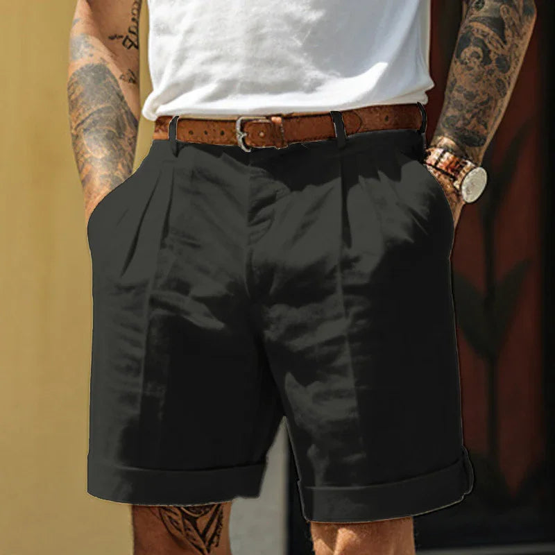 Man wearing black pleated shorts with brown belt, white shirt, and tattooed arms