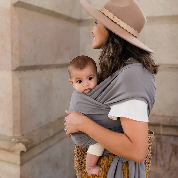 Woman wearing beige hat carrying baby in gray wrap sling against stone wall