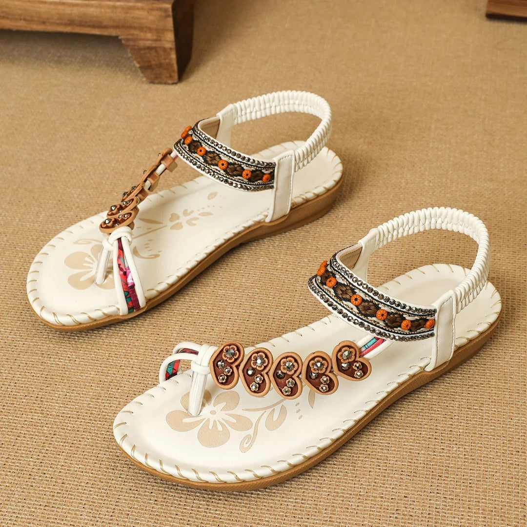 Pair of white bohemian sandals with floral embellishments and braided ankle straps on textured surface