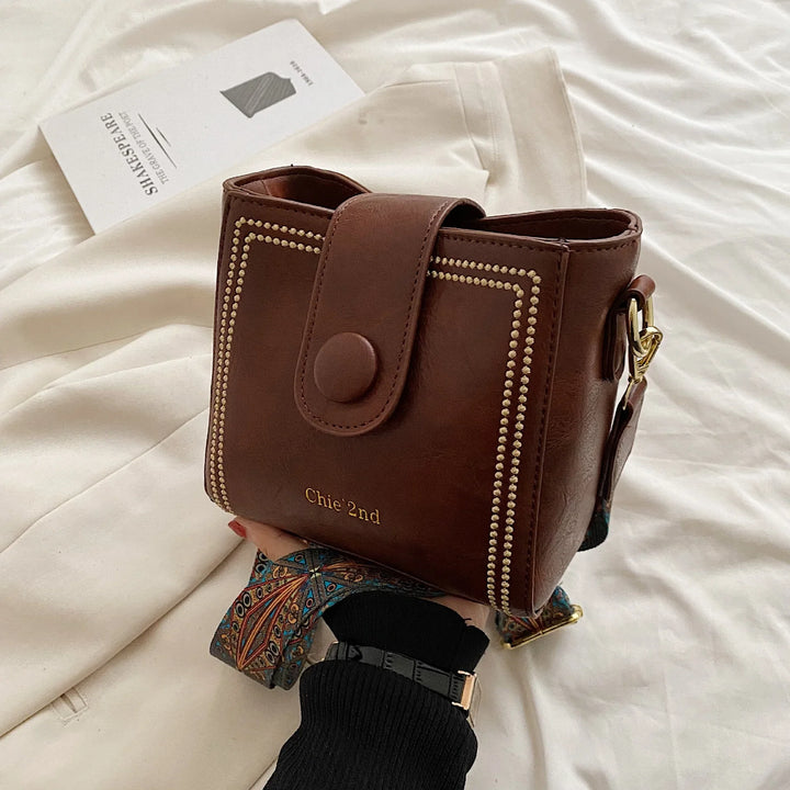 Hand holding brown leather Chie 2nd crossbody bag with decorative stitching and patterned strap on white fabric