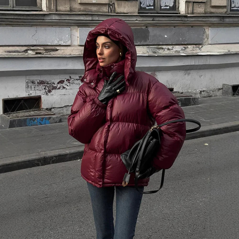 Woman wearing a hooded burgundy puffer jacket, black leather gloves, holding a black handbag on a city street