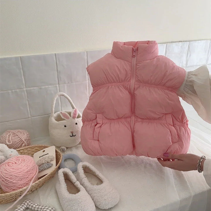 Pink puffer vest for baby on white tiled background with soft slippers, yarn, and bunny basket