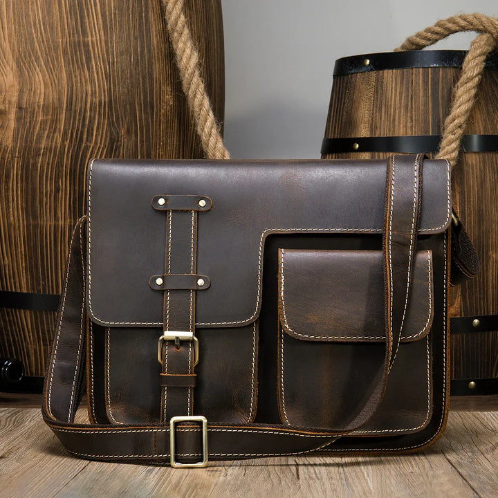 dark brown leather messenger bag with front buckle pocket placed on wooden floor with barrels background