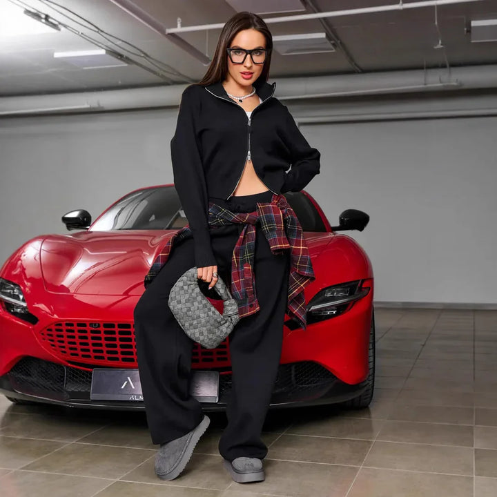 Young woman in black loungewear with plaid shirt and gray handbag posing in front of red sports car in garage