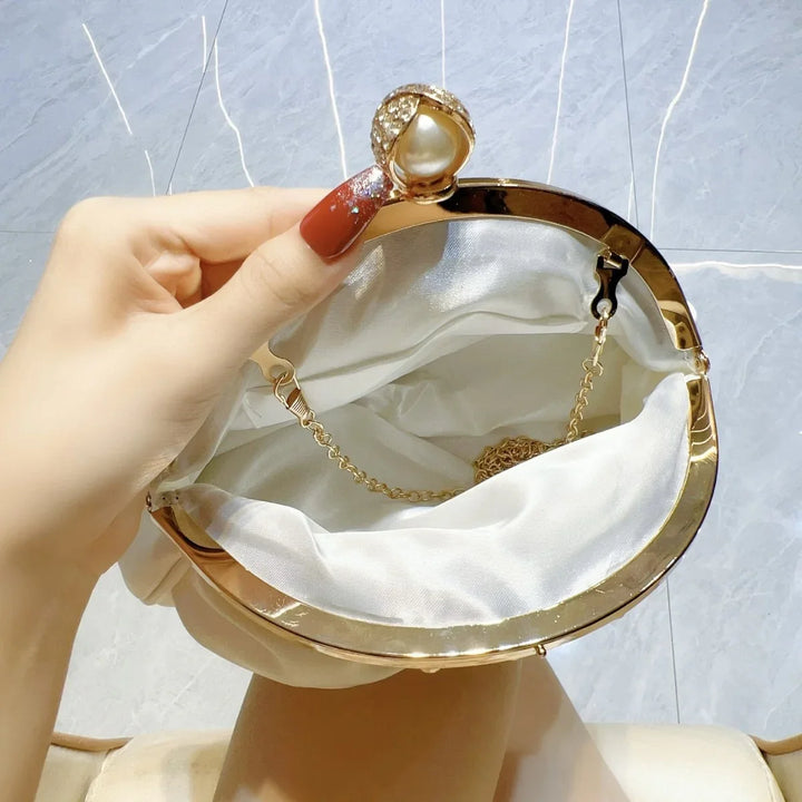 Hand holding open a white satin purse with gold frame and pearl clasp, showing inside chain strap