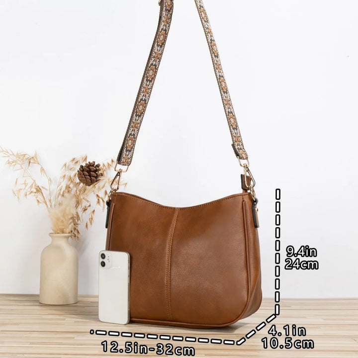 Brown leather shoulder bag with patterned strap, next to white smartphone and vase with dried flowers
