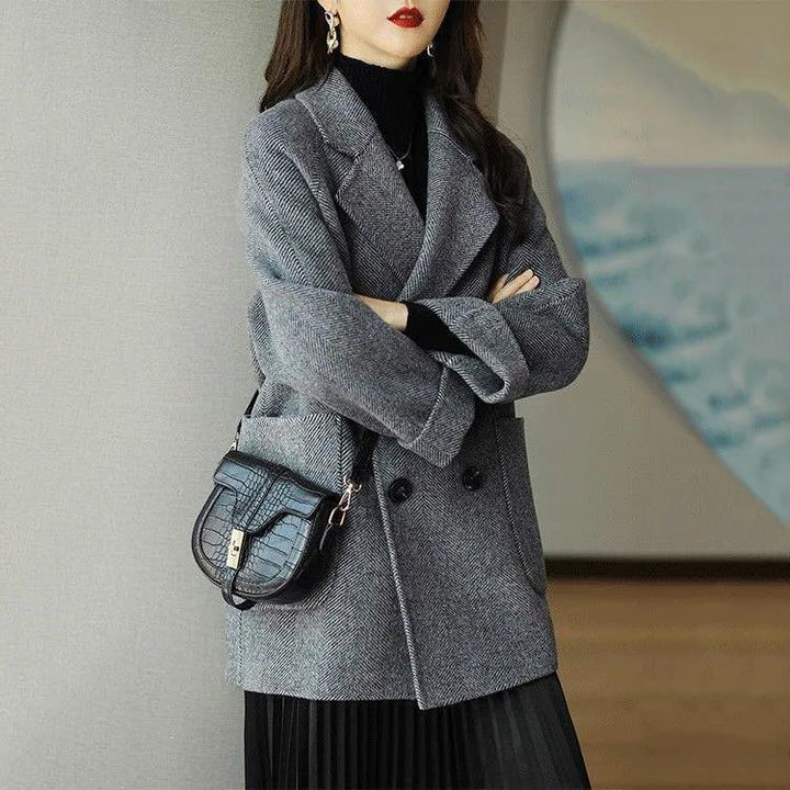 Woman wearing gray herringbone double-breasted coat with black pleated skirt and black leather handbag