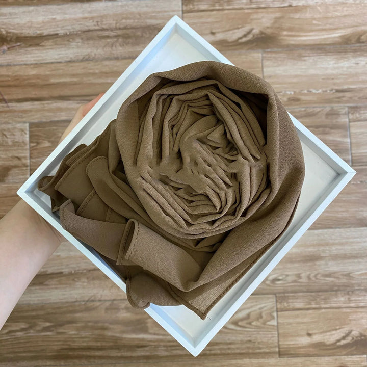 Hand holding white square tray with neatly rolled brown fabric scarf on wooden floor