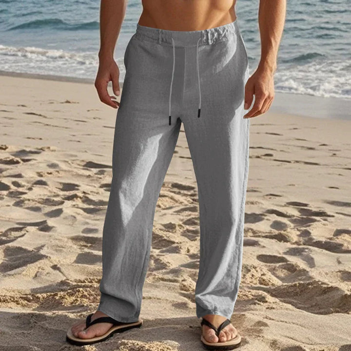 Man wearing light gray drawstring beach pants and flip-flops standing on sandy ocean beach