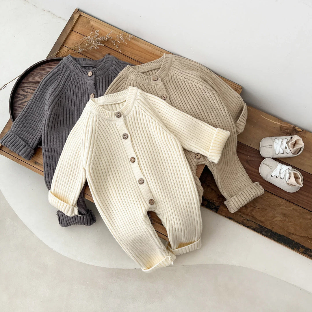 Three ribbed knit baby rompers in cream, beige, and gray with wooden buttons, displayed on wooden surface
