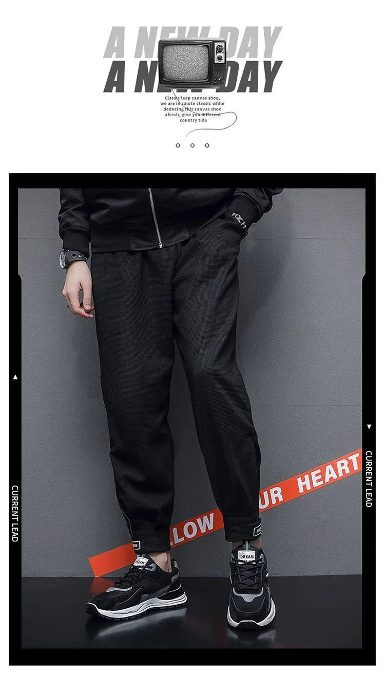 Man wearing black jogger pants and black white sneakers standing against gray wall with red ribbon