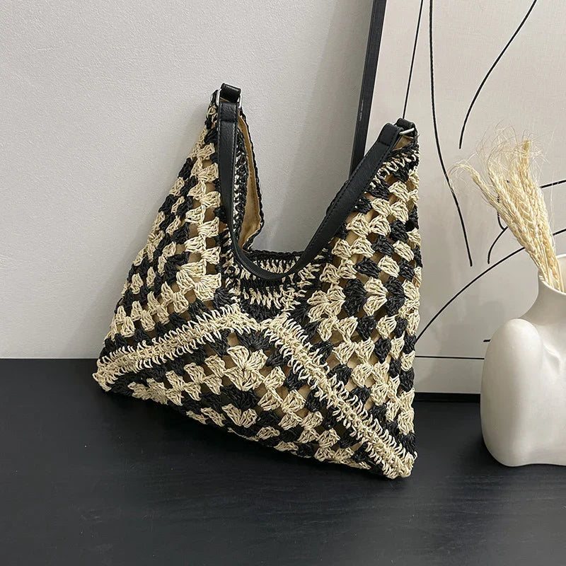 Black and beige crochet handbag with black leather straps on black surface beside modern vase with dried grass