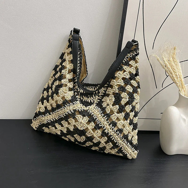 Black and beige crochet handbag with black leather straps on black surface beside modern vase with dried grass