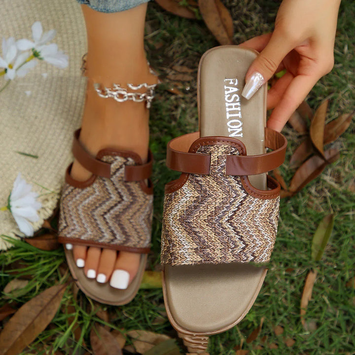 Woven brown slip-on sandals with leather straps, one worn on foot with white nail polish