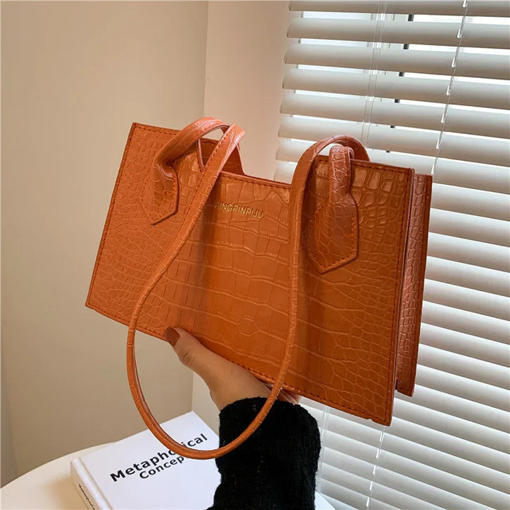 Hand holding orange crocodile texture leather handbag near window blinds and white book