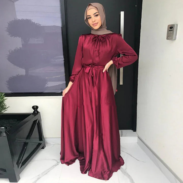 Woman wearing elegant maroon satin long dress and light brown hijab standing indoors near black door