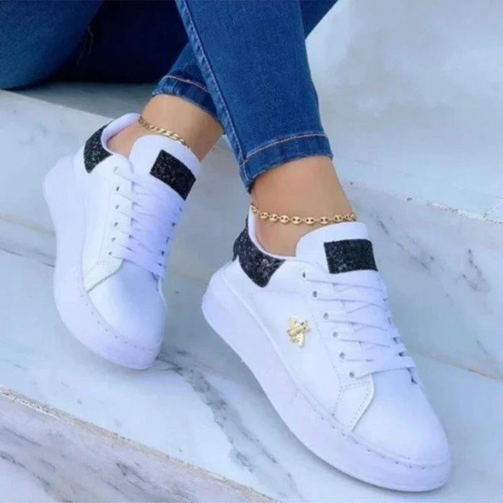 White sneakers with black glitter heel patches and gold bee detail worn with blue jeans and gold anklet on marble steps