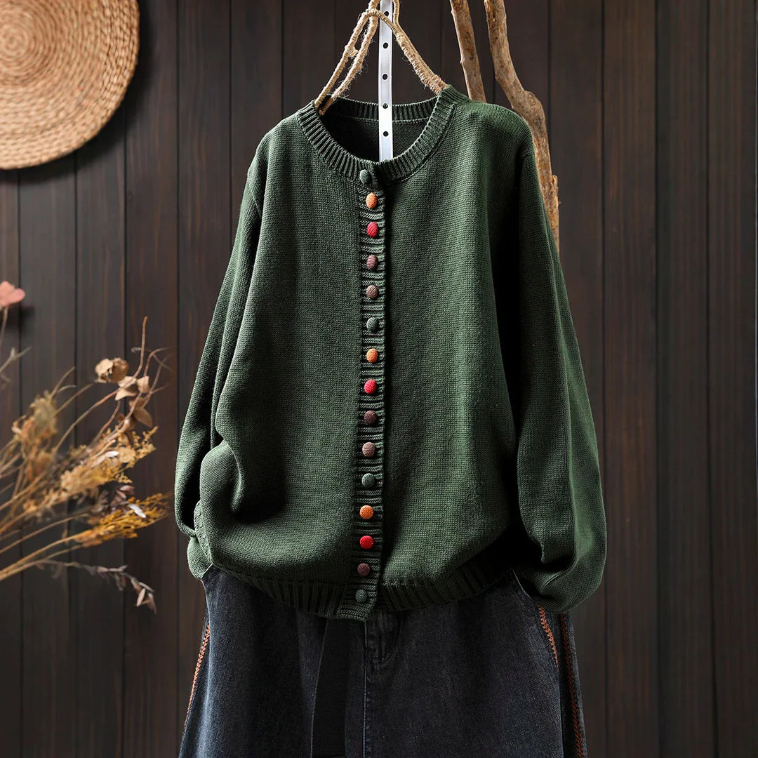 Dark green knit cardigan with colorful round buttons hanging on rustic branch against wooden wall