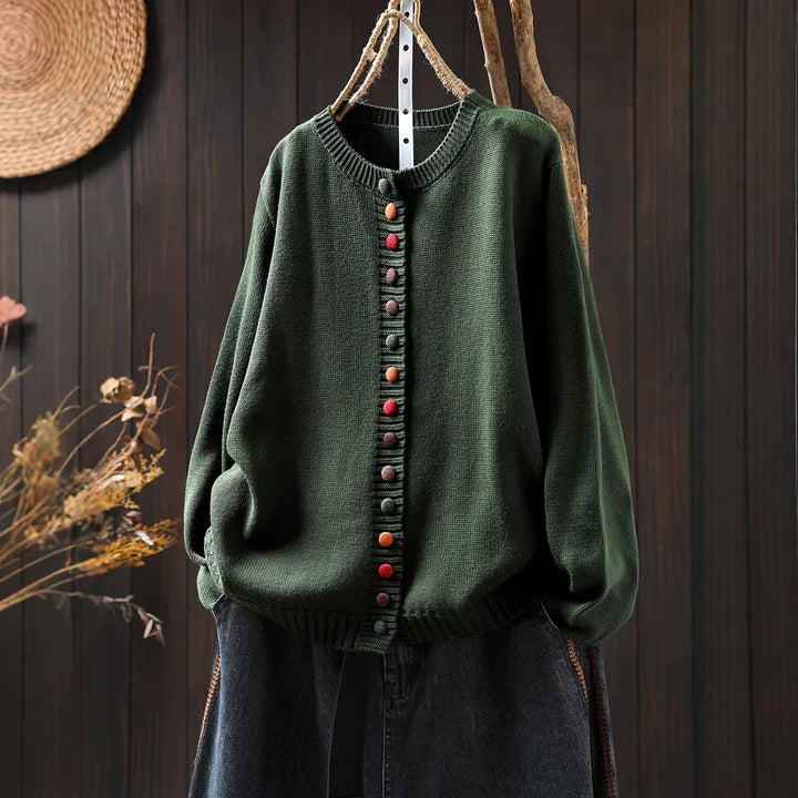 Dark green knit cardigan with colorful round buttons hanging on rustic branch against wooden wall
