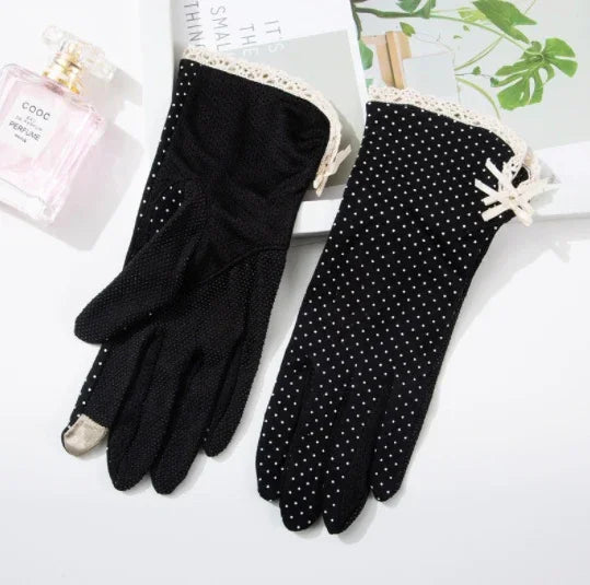 pair of black polka dot gloves with lace trim and bow detail displayed on white surface with perfume bottle and green leaves