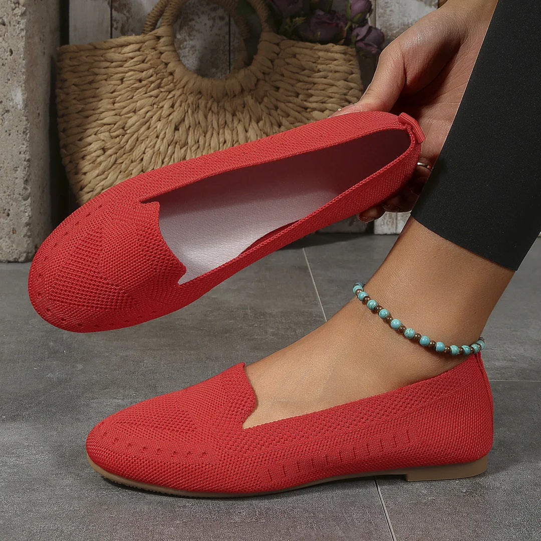 Red knit flat shoes worn on foot and held in hand with a beaded anklet on gray tile floor