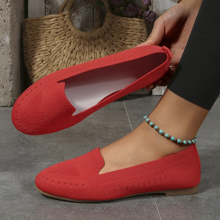 Red knit flat shoes worn on foot and held in hand with a beaded anklet on gray tile floor