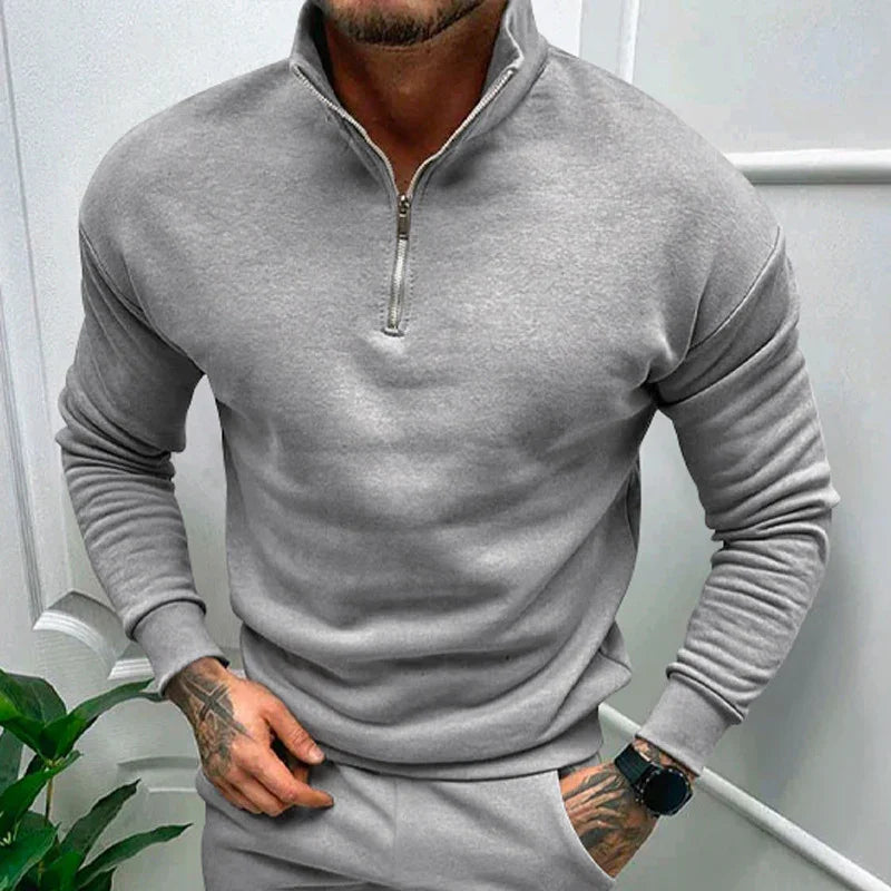 Man wearing grey quarter-zip sweatshirt and matching pants with tattoos on hand and wristwatch