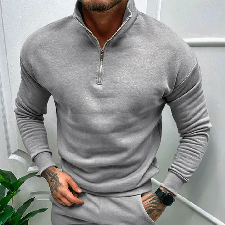 Man wearing grey quarter-zip sweatshirt and matching pants with tattoos on hand and wristwatch