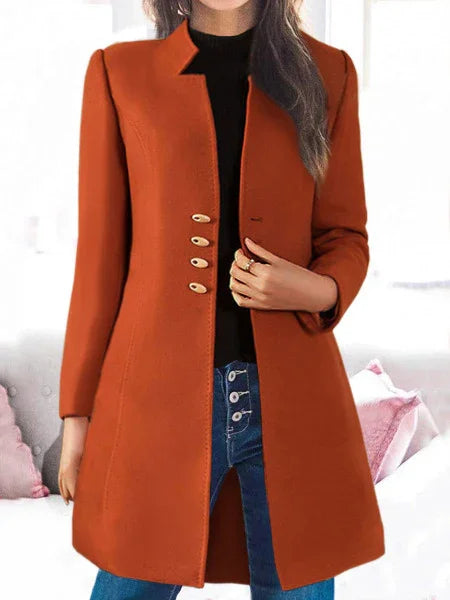 Woman wearing a stylish rust orange long coat with gold buttons, black top, and blue jeans indoors