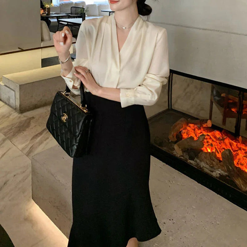 Woman in elegant cream blouse and black skirt holding black quilted handbag near modern fireplace indoors