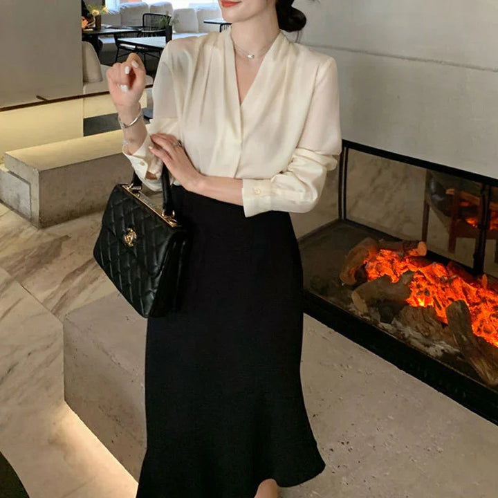 Woman in elegant cream blouse and black skirt holding black quilted handbag near modern fireplace indoors