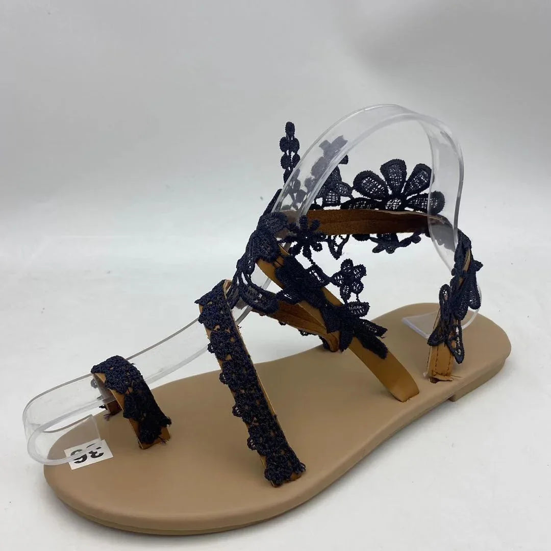 Tan flat sandal with black lace floral straps displayed on a clear foot model
