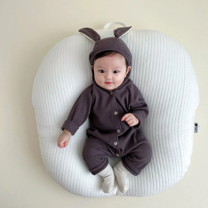 Baby wearing brown ribbed bunny ear onesie sitting on white cushioned pillow against beige background