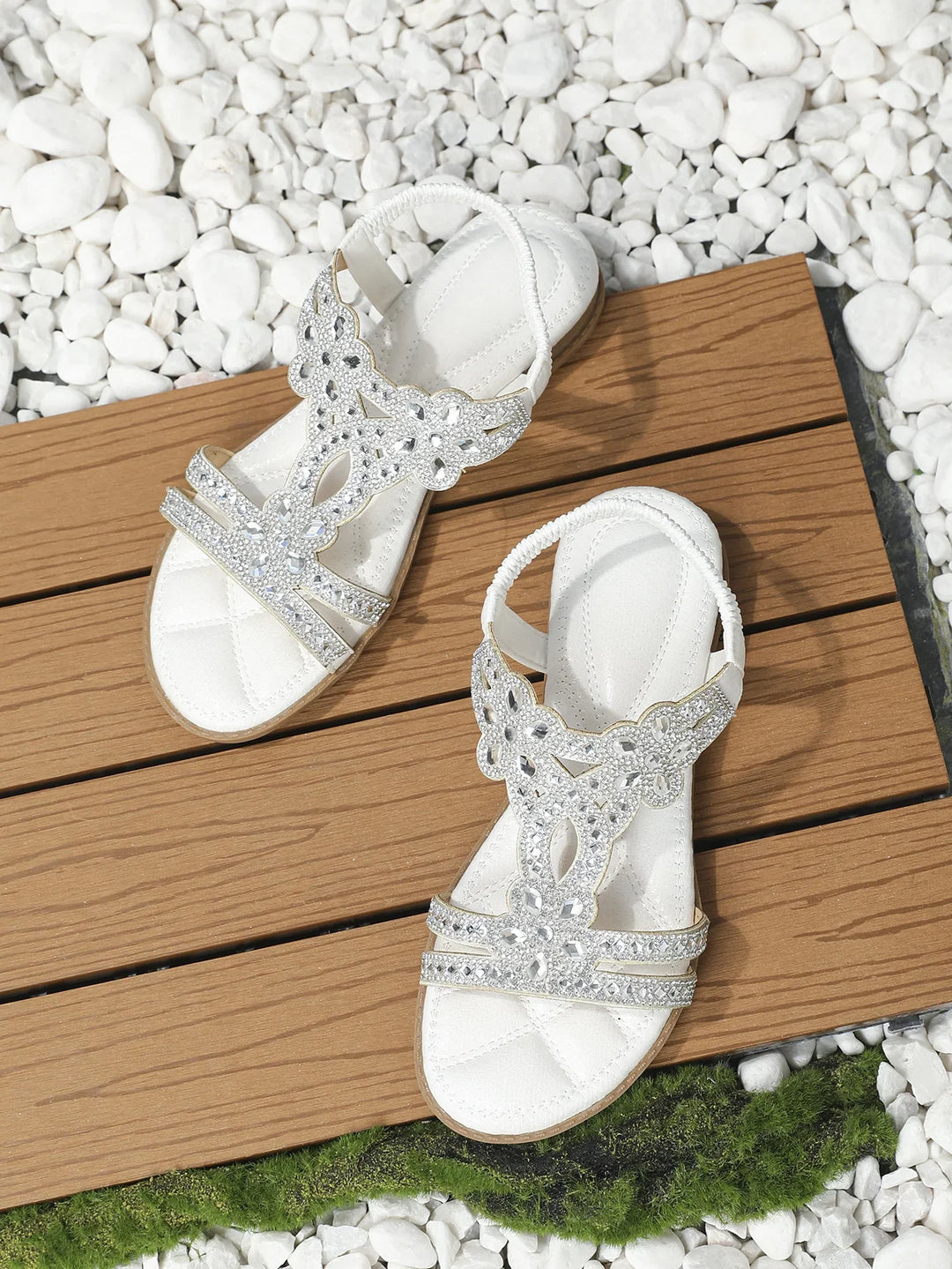 White sandals with rhinestone embellishments on wooden planks surrounded by white stones and green moss