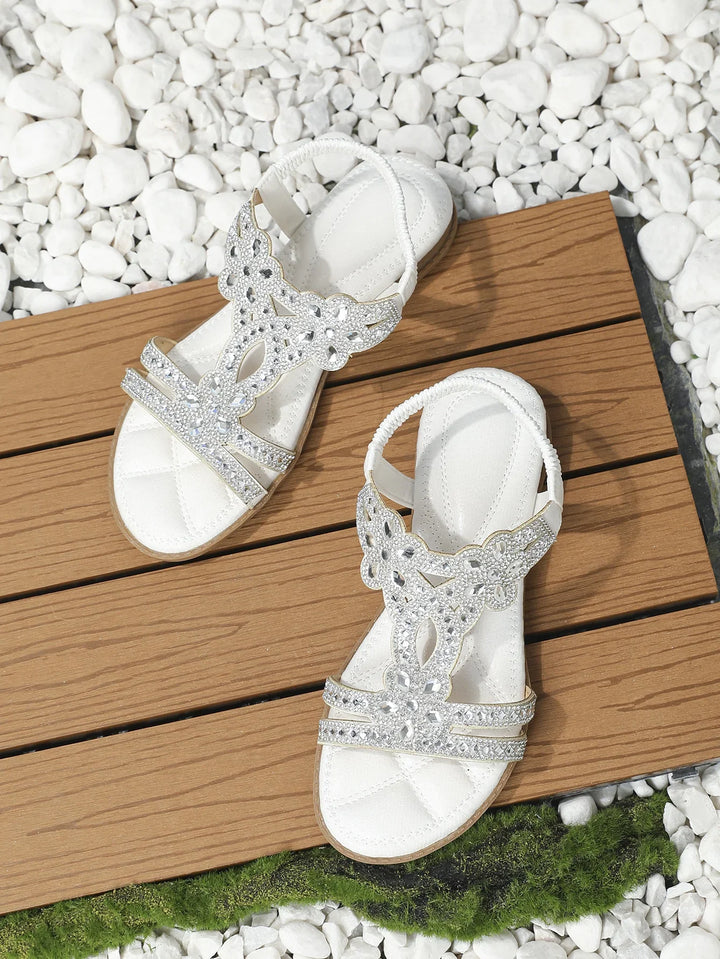 White sandals with rhinestone embellishments on wooden planks surrounded by white stones and green moss
