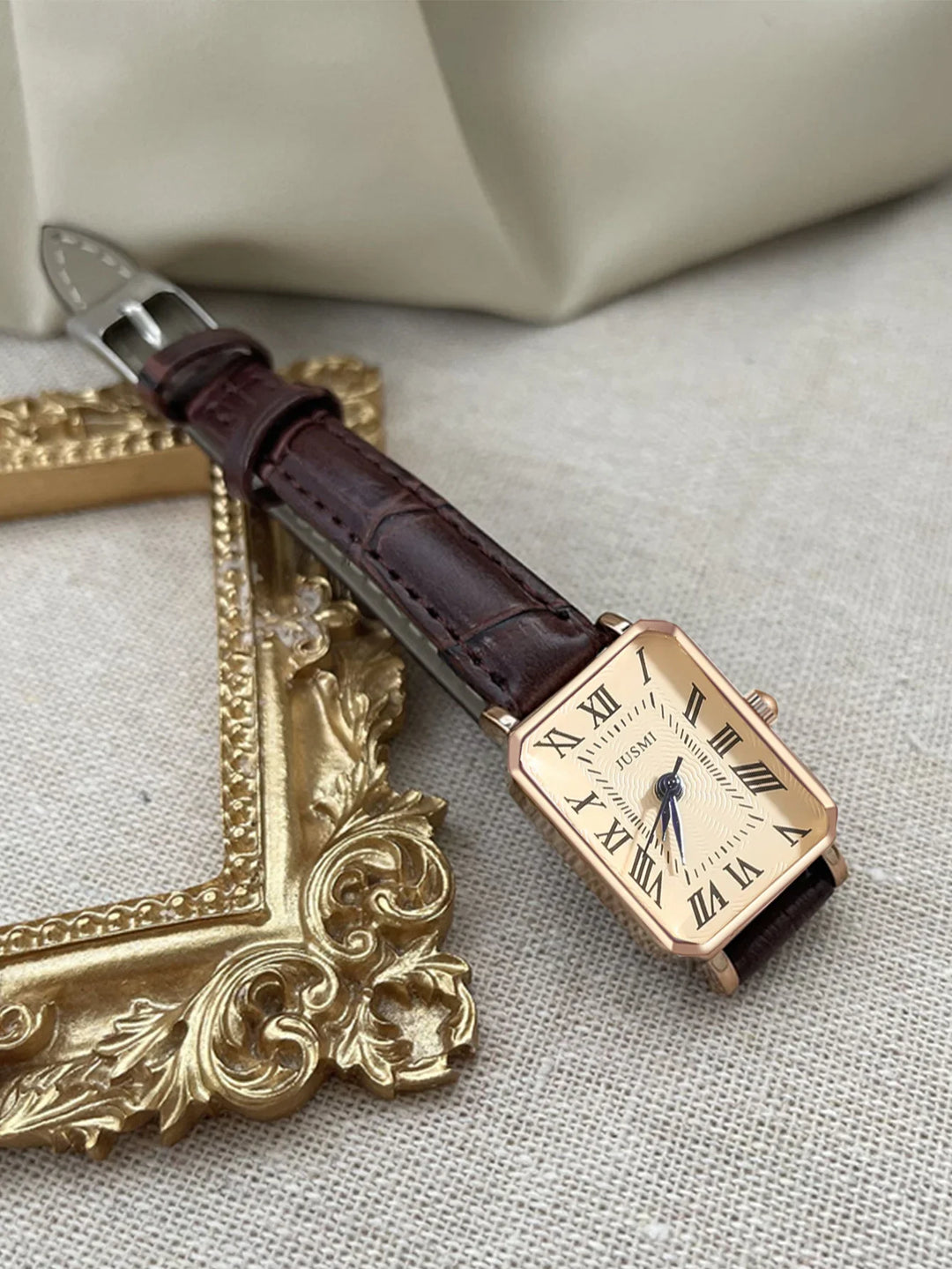 Rectangular gold wristwatch with Roman numerals and dark brown leather strap on beige fabric near ornate gold frame
