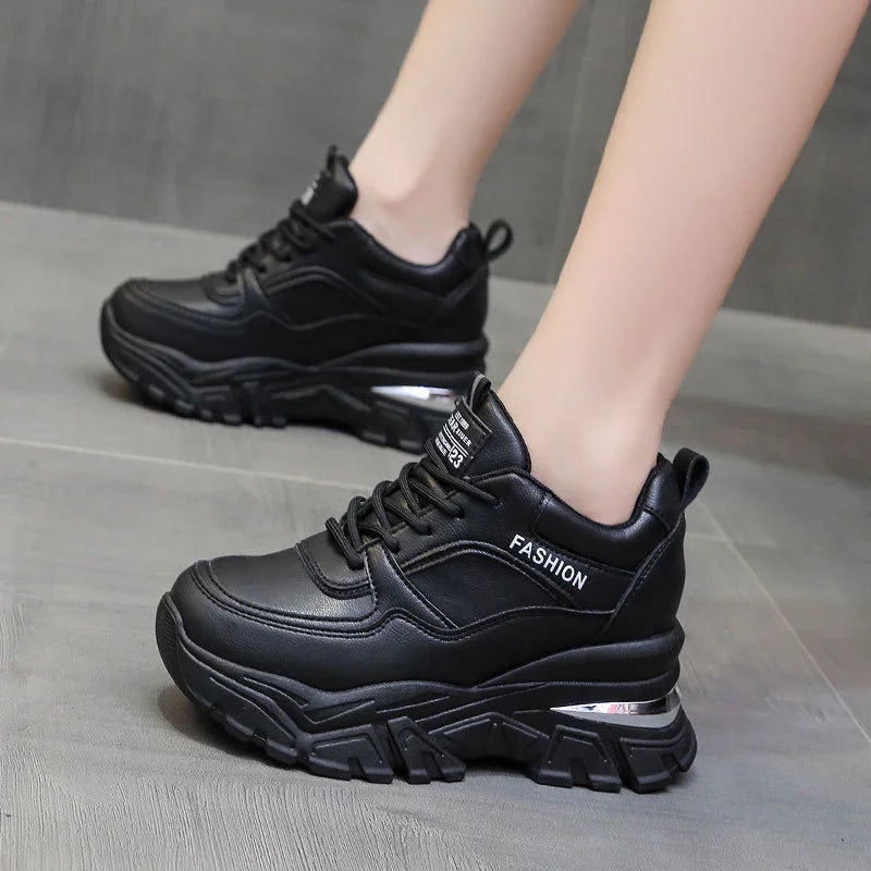 Black chunky platform sneakers with thick soles and fashion text on side worn on gray floor