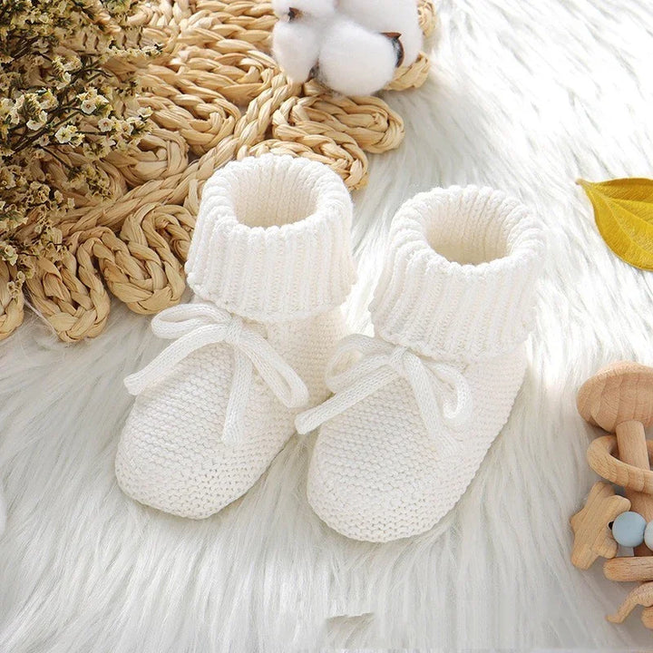 Pair of white knitted baby booties with ties on a soft white fur surface surrounded by cotton and wooden toys