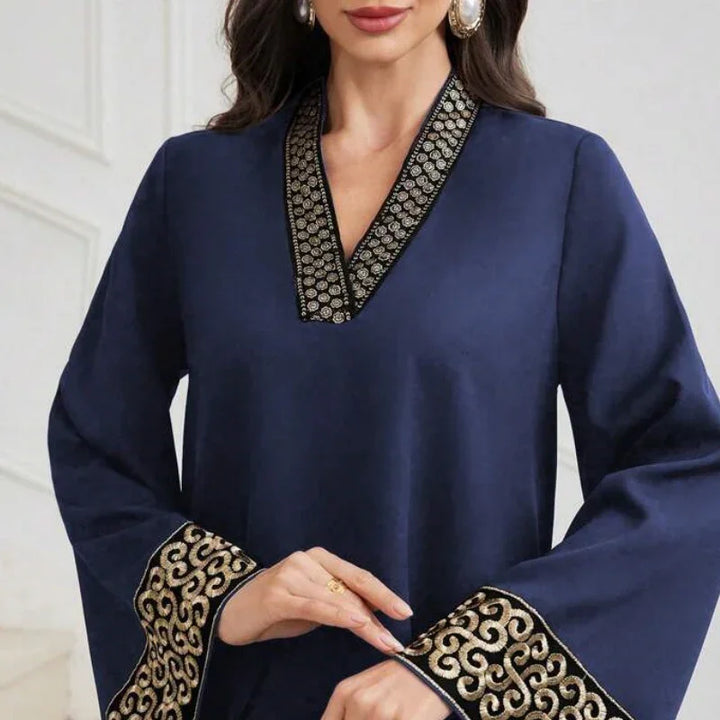 Woman wearing navy blue dress with embroidered gold and black V-neck and wide cuffs, posing indoors